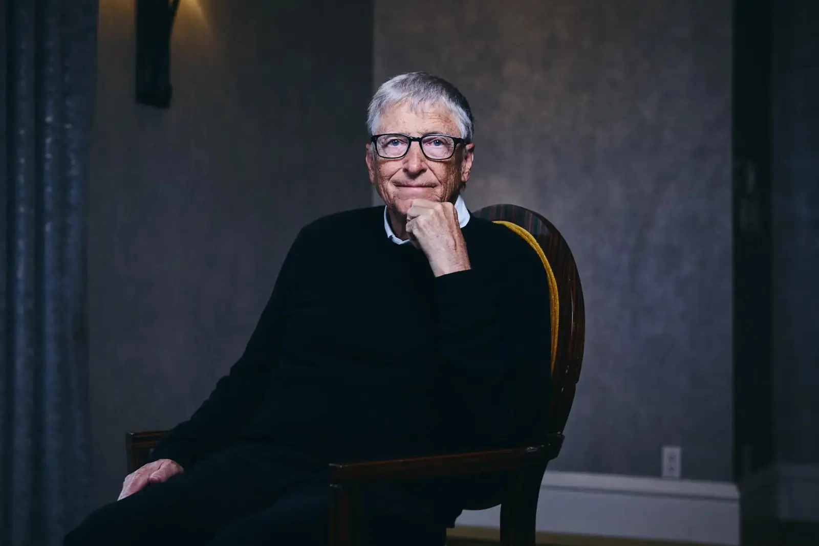 بیل گیتس (Bill Gates) با عینک در حالی که روی صندلی نشسته است